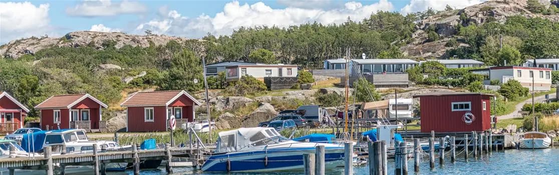 Bild av Rörviks camping: En naturskön kuststräcka med en liten by vid havet. I förgrunden syns en båt vid en brygga, och en vik omgiven av grönskande öar.