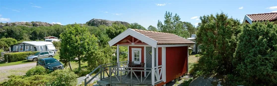 En timmerstuga omgiven av en välskött trädgård på Rörviks camping, med en gräsmatta och träd i bakgrunden. 