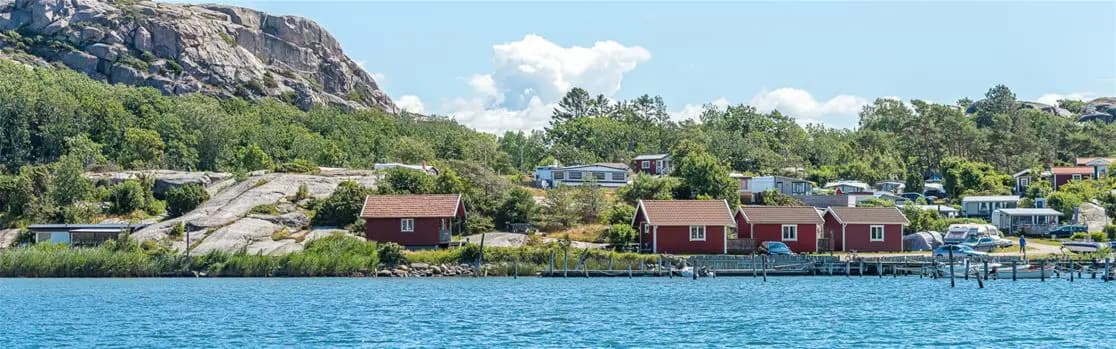 En naturskön vy över Rörviks camping, med en stuga vid kanten av ett lugnt vattendrag och omgivande kustlandskap.