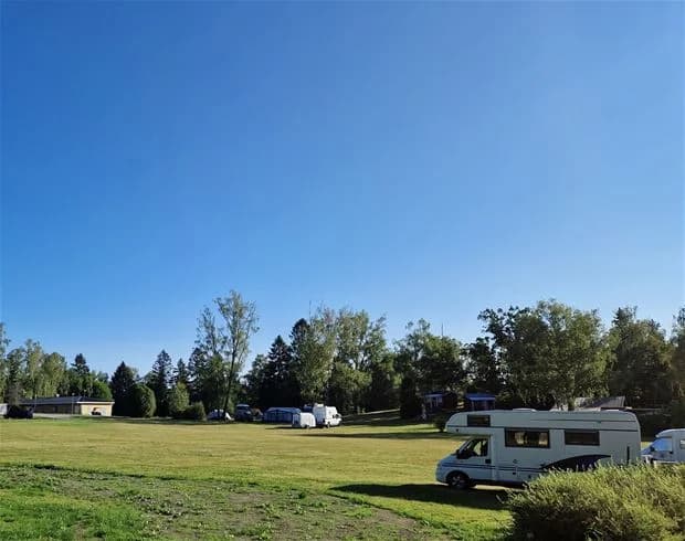 En husbil är parkerad på ett grönskande campingområde omgiven av träd och blå himmel, med en rymlig gräsmatta i förgrunden.