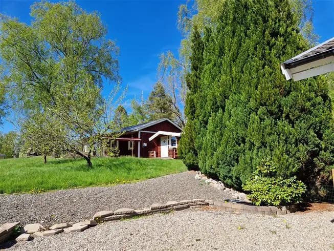 En lummig campingplats med en vit stuga omgiven av välskötta buskar, gräsmatta och trädgård. Barrträd skapar en inramning runt byggnaden.
