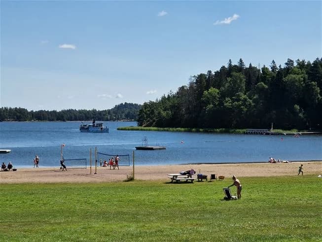 "En bild från Farstanäs camping & havsbad som visar en strand vid kusten med vatten i bakgrunden. Människor ses njuta av fritidsaktiviteter vid sjön, med båtar som flyter på vattnet och en kustlinje som sträcker sig längs horisonten."