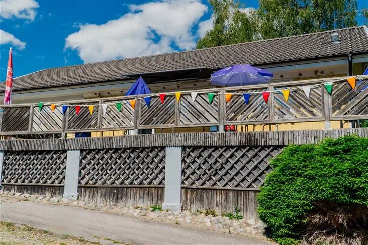 Bild av en del av Farstanäs Camping & Havsbad. Föreställer ett campingområde med staket, skuggiga träd, veranda och ett staket som markerar tomtgränsen. Soligt väder bidrar till en avslappnad atmosfär.