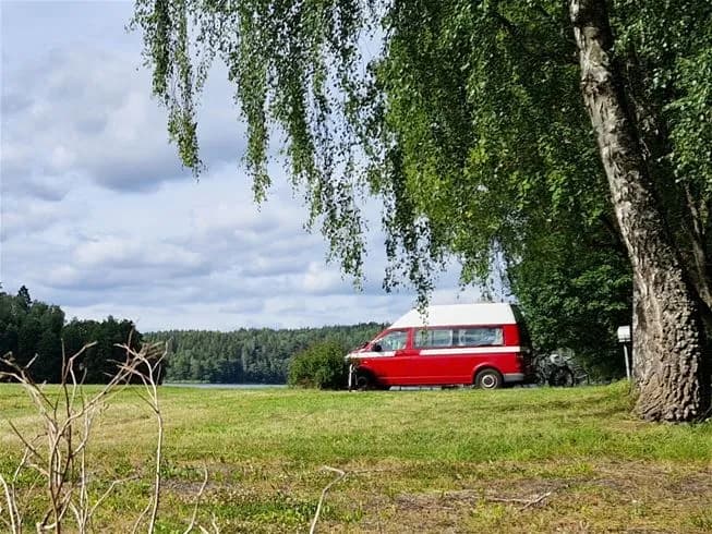 En vit Volkswagen-minibuss står parkerad på Farstanäs Camping & Havsbad, omgiven av grönska och himmel i bakgrunden.