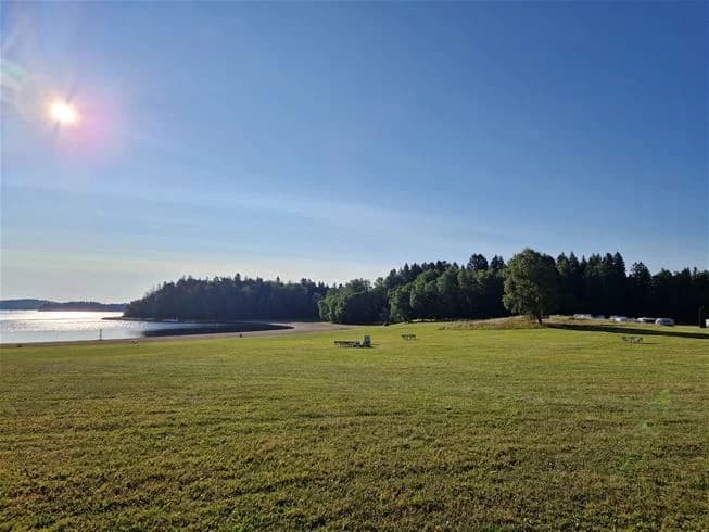 En vidsträckt grön gräsäng sträcker sig mot horisonten, med en glimt av sjöns glittrande yta och en sandig strandlinje i bakgrunden, vid Farstanäs camping & havsbad.