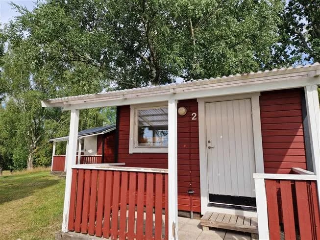 En trästuga med ett sluttande tak och dörren lätt öppen, omgiven av gröna omgivningar på Farstanäs camping & havsbad.