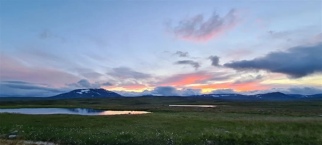 En vy över Jormvattnets Fiskecamp med en storslagen natur i bakgrunden, omgiven av molntäckt himmel, bergslandskap och gröna kullar.