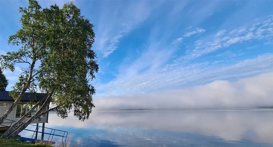 En stilla sjö omgiven av skogsklädda berg, spegelblankt vatten reflekterar moln på himlen, morgondimma svävar över sjön vid Jormvattnets fiskecamp.