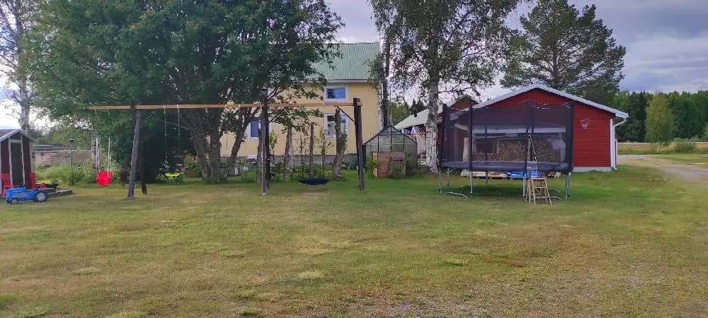 En röd stuga med vita knutar och brunt tak, omgiven av en välklippt gräsmatta och fåtöljgrupper, belägen i ett lantligt område under en klar himmel.