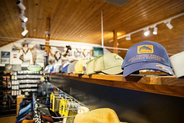 En man med keps och aktivitets-tröja står vid en hylla med campingvaror. Bakom honom syns logotyper och utrustning för friluftsliv.