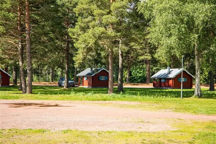 Bild av Älvdalens camping med en grön gräsmatta och omgivande träd. I bakgrunden syns stugor och små hus i ett lantligt område.