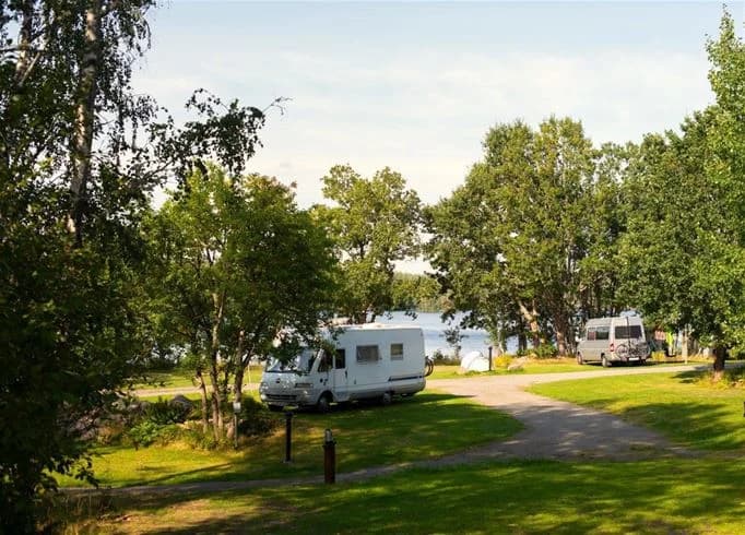 En husvagn parkerad vid stranden i ett naturskönt campingområde vid Långsjön, omgiven av grönska och en glittrande sjö i bakgrunden.
