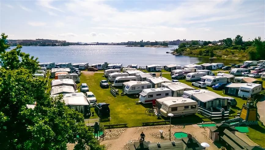 Flygbild över Dragsö Camping & Stugby med kustlinje, parkeringsplats, strand och flera fordon som lastbilar och skåpbilar. Det omgivande området visar en vacker resortort vid en bukt.