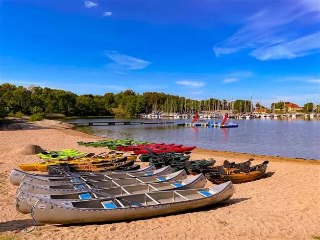 En bild på Dragsö Camping & Stugby med båtar förtöjda vid en brygga i en naturskön sjömiljö. Vattnet sträcker sig ut mot horisonten, omgiven av grönskande skog och kustlinje, vilket inbjuder till rekreation och avkoppling i naturen.