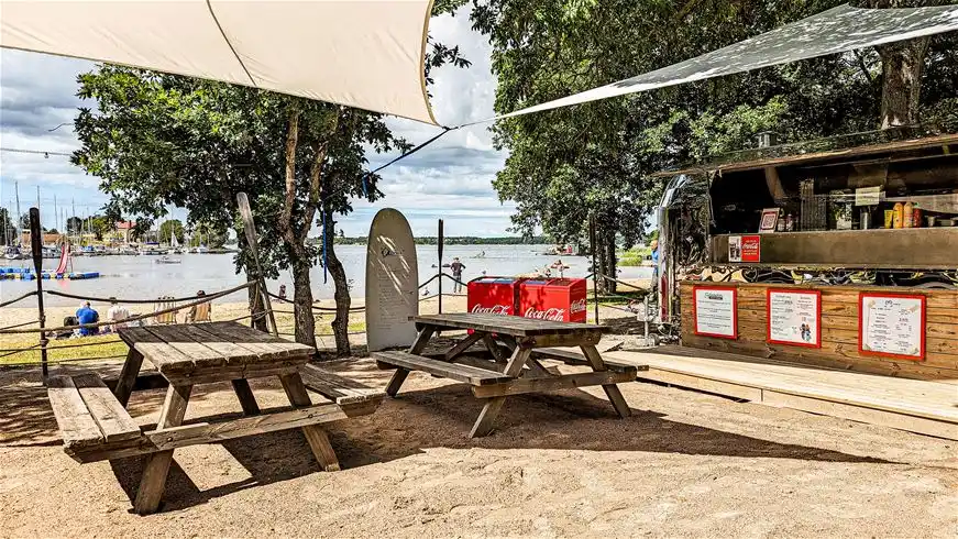 En solig dag vid Dragsö Camping & Stugby med picknickbord och bänkar under träden, omgivna av sand och parkmiljö.