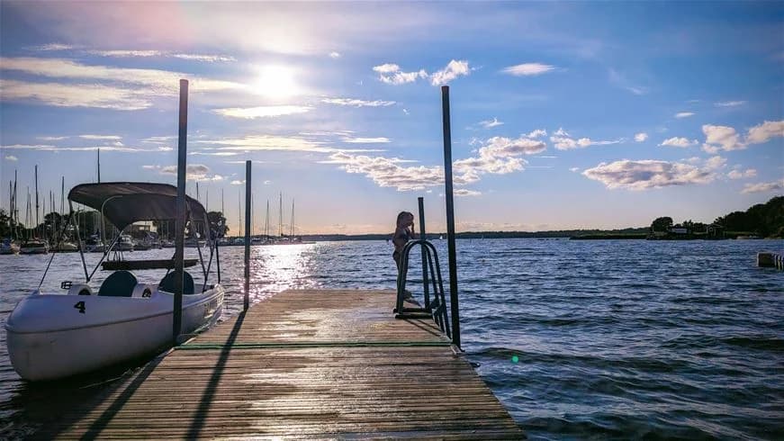 En solig morgonvy över Dragsö camping & stugby med blått vatten i förgrunden, en båt vid bryggan och en klar horisont i bakgrunden.
