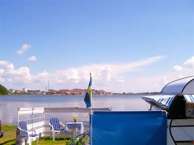 Bild av Dragsö Camping & Stugby som visar en sjö med flera båtar förtöjda vid en brygga. Utemöbler står på gräset i förgrunden, omgivna av skuggade träd.