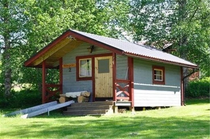 En trästuga med sadeltak och dörr, omgiven av en lantlig miljö.