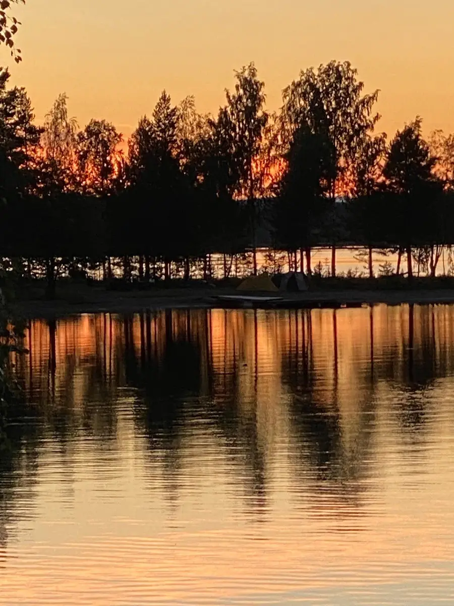 En naturskön vy över en sjö vid skymning med solnedgångens varma färger som reflekteras i vattnet, omgiven av grönskande natur i kojbyns camping.