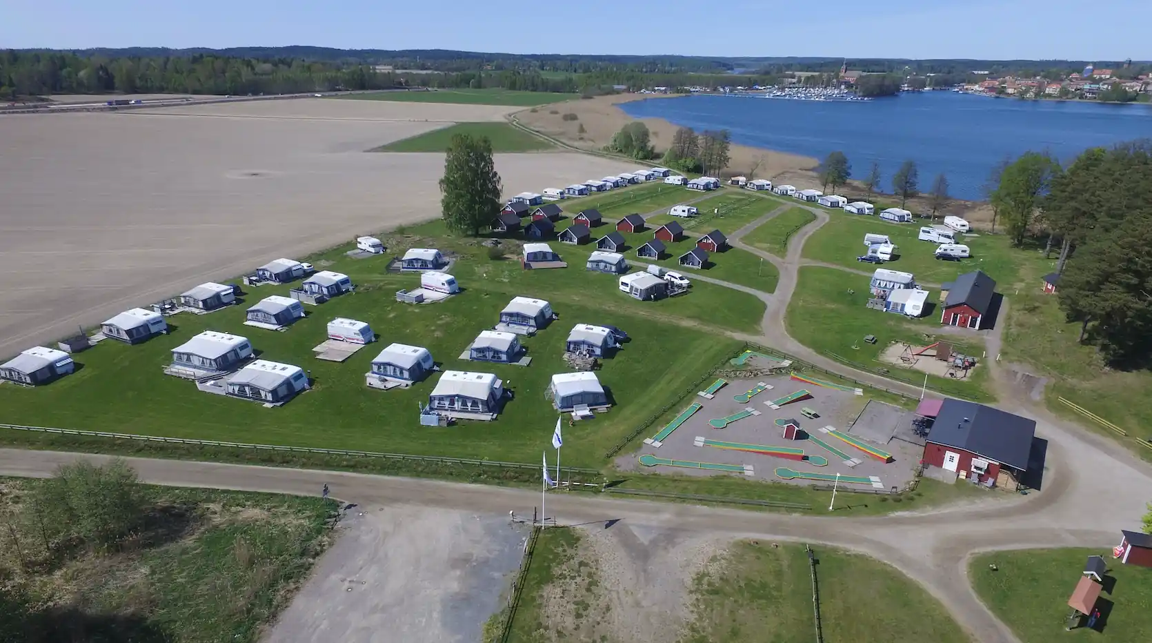 Flygfoto över Husabergsudde camping som visar en park med husbilar parkerade på campingplatser. I bakgrunden syns en marina vid vattnet och flera fordon, inklusive långtradare, står uppställda på parkeringsområdet.