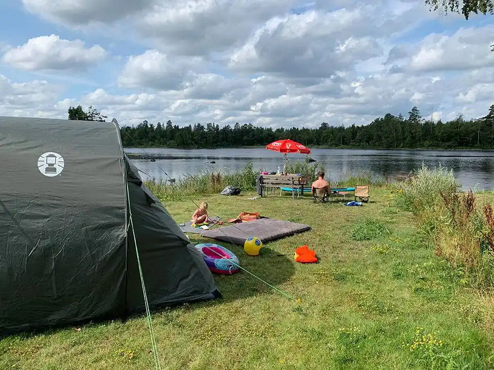 Bilden visar Alsterbro camping med tält uppställda nära en sjö. Området har en naturlig miljö med gräsbevuxna stränder och vatten i bakgrunden, vilket ger en idyllisk plats för rekreation och avkoppling.