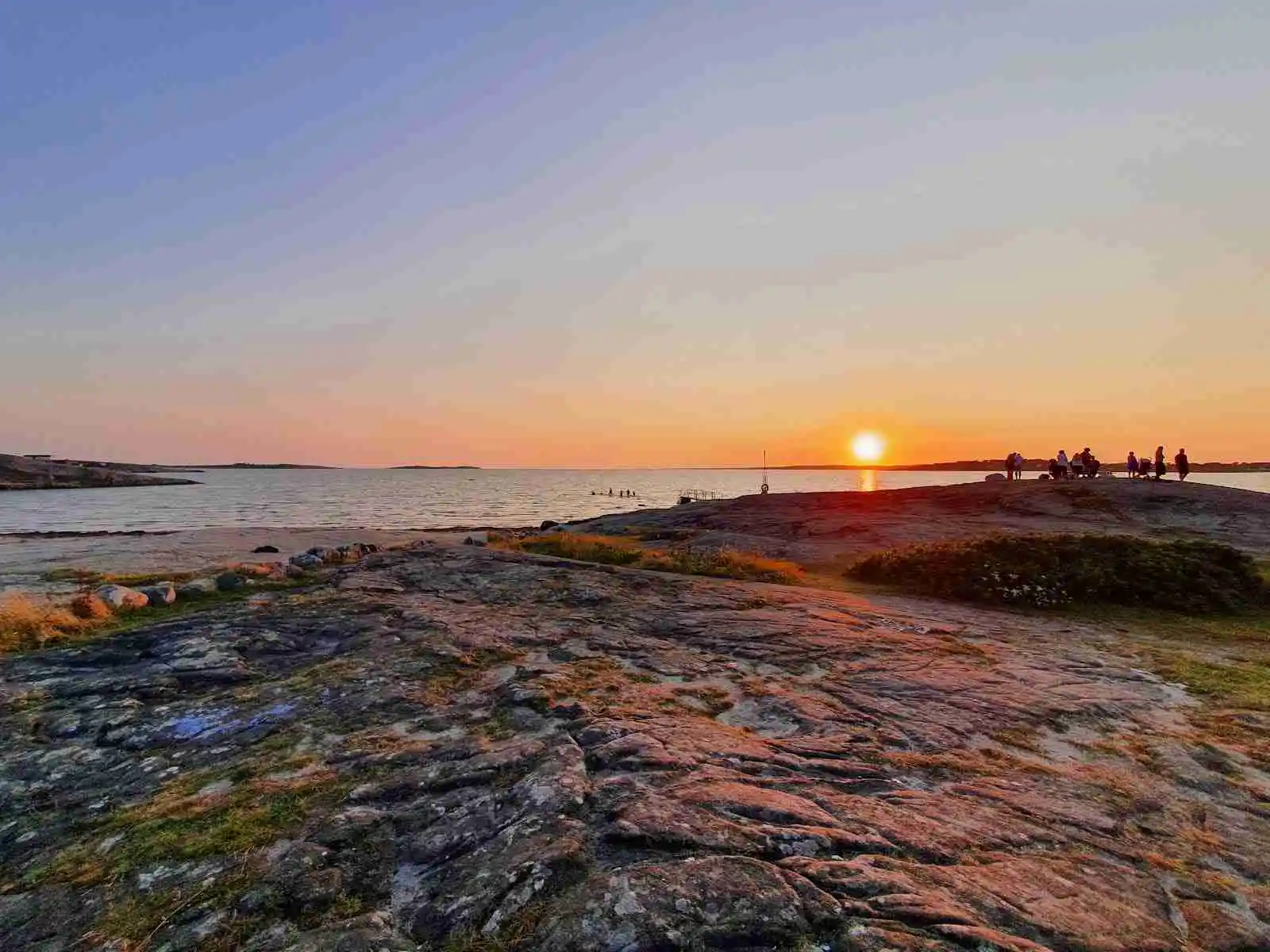 En naturskön vy över kustlinjen vid Vallersvik camping och vandrarhem, med vatten som möter horisonten under en vacker solnedgång. Klippor skymtar i förgrunden och himlen glöder i varma skymningsfärger.