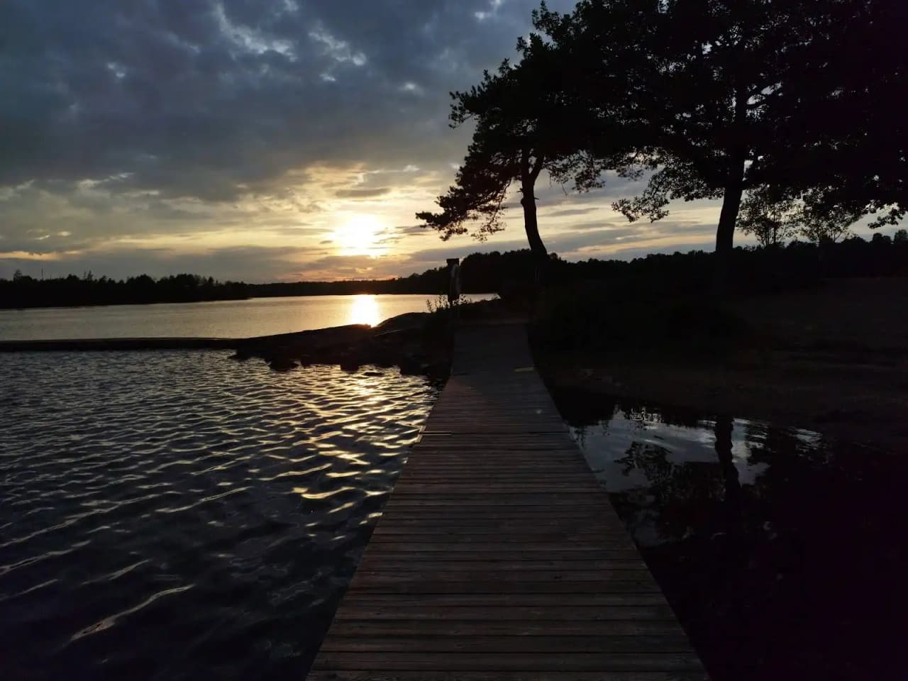 En stilla sjö vid solnedgången på Alsterbro camping, med en klar himmel och ljusrosa och orange nyanser i horisonten, omgiven av natur.