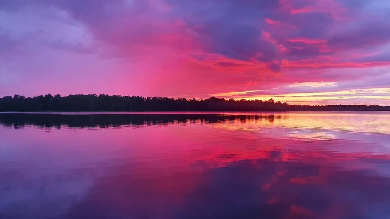 En solnedgång över en sjö, med moln som reflekteras i det stilla vattnet. Himlen är färgad i blått och rött, vilket skapar en rogivande atmosfär.