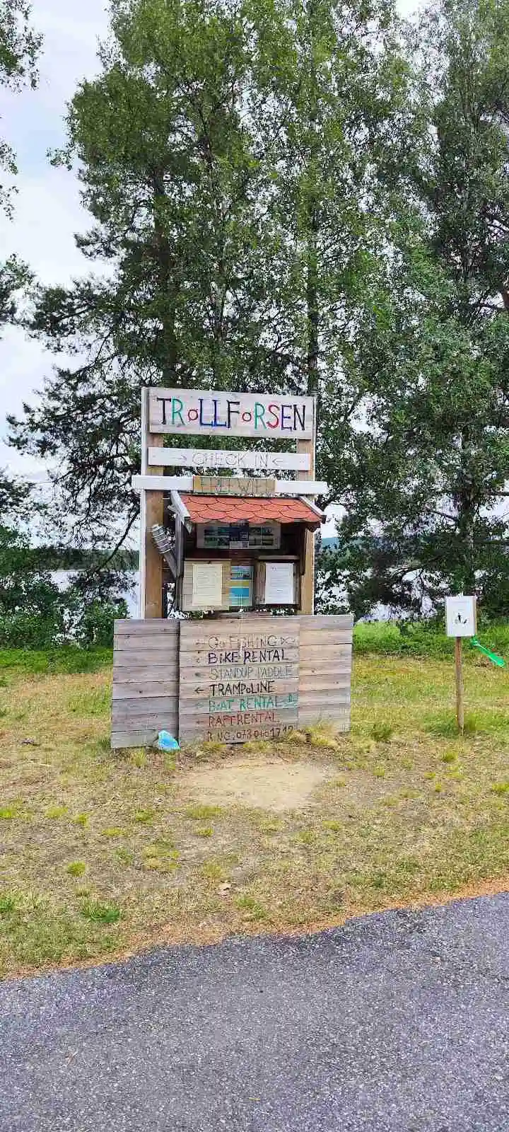 En träskylt vid ingången till Trollforsen Camping & Stugor med omgivande grönska och en grusad gångväg i bakgrunden. En mindre träbyggnad syns på avstånd.