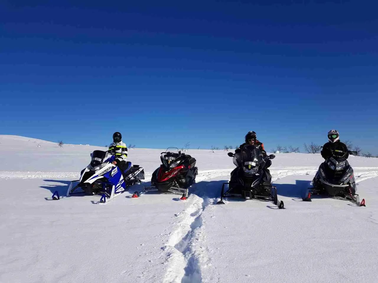 En person kör snöskoter genom ett snötäckt landskap vid Grönfjälls Fritidsby, iklädd vinterkläder och hjälm. Det omgivande området är vitt av snö och bjuder in till vinteraktiviteter och äventyr.