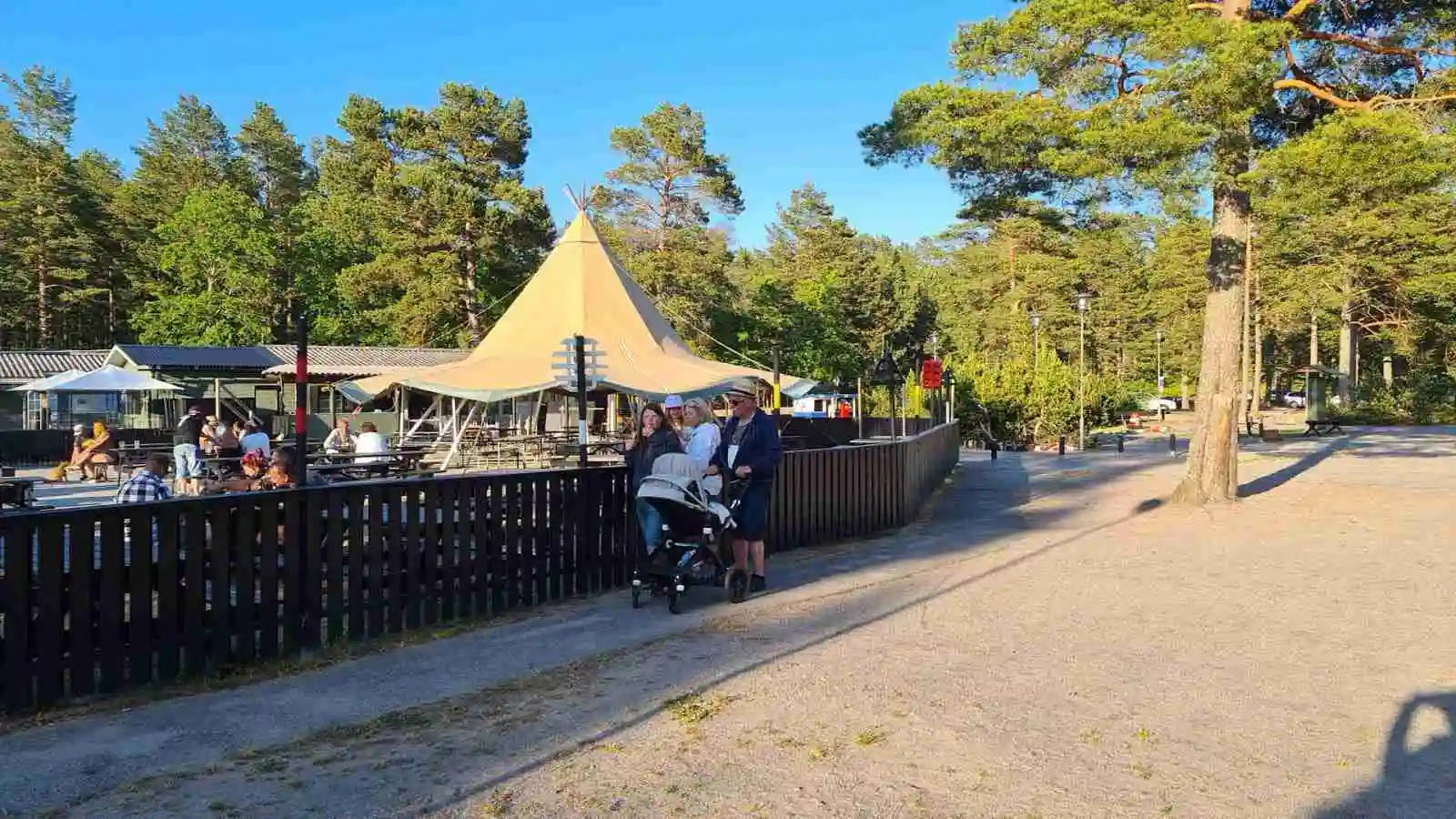En solig dag vid Stenö havsbad & camping med utemöbler som inkluderar bänkar och picknickbord under skuggande träd. En inbjudande miljö för rekreation och avkoppling i det fria.