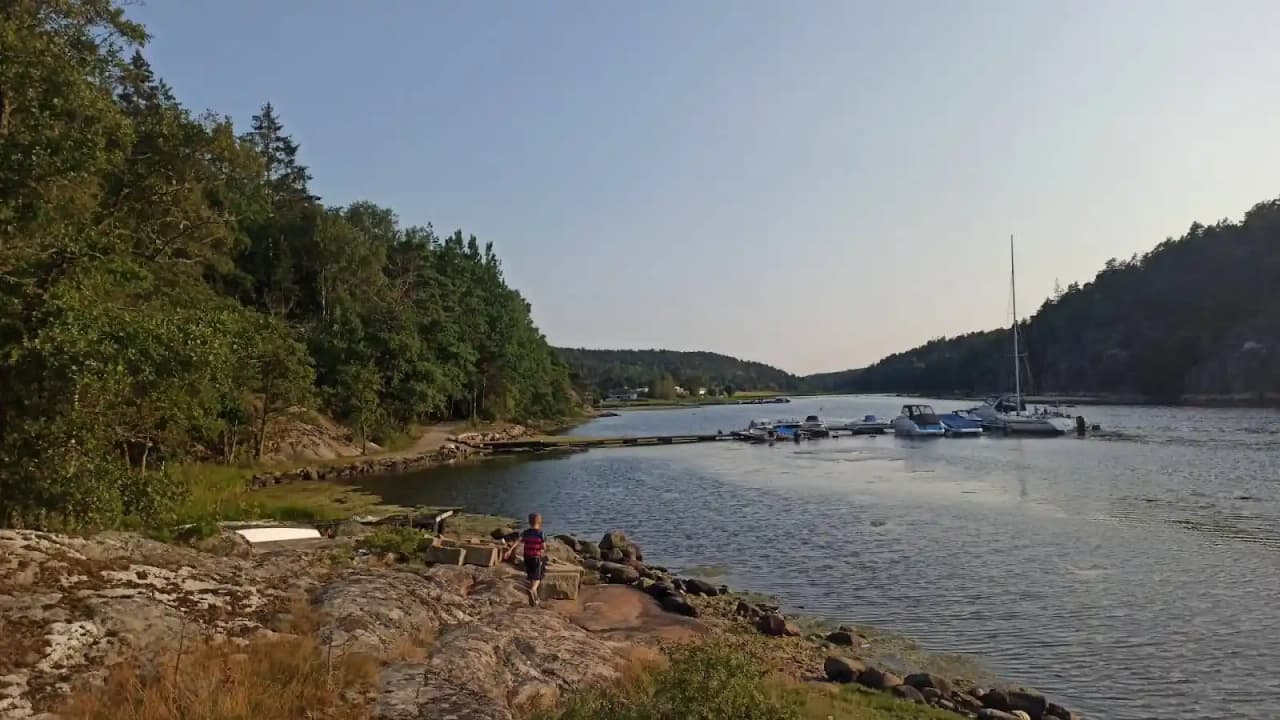 En vy över Ylserød camping med en båtramp vid vattnet. En liten båt ligger förtöjd vid strandkanten, omgiven av grönskande landskap och ett glittrande vattendrag.