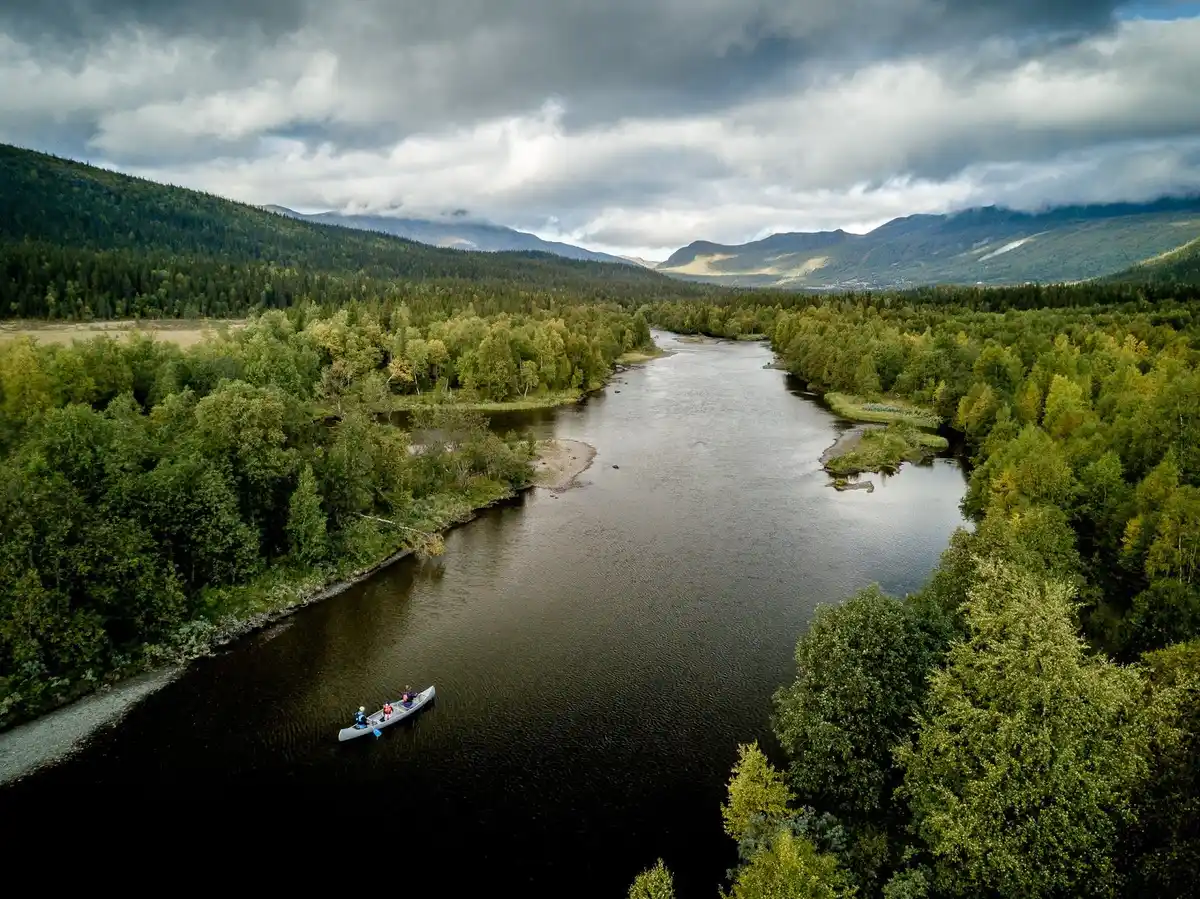 En naturskön vy vid Camp Kittelfjäll med en glittrande vattenyta i förgrunden och majestätiska berg täckta av moln i bakgrunden.