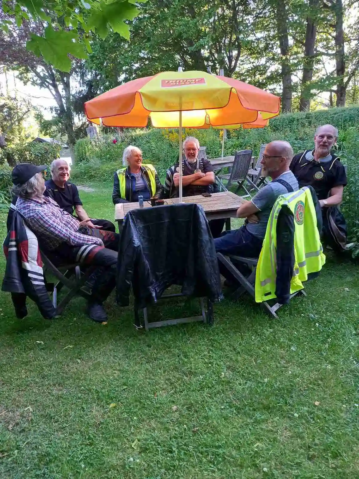 En campingplats med utomhusmöbler, inklusive bord och stolar, och ett parasoll för skugga. Människor njuter av fritiden och sällskap i en gemytlig utomhusmiljö.