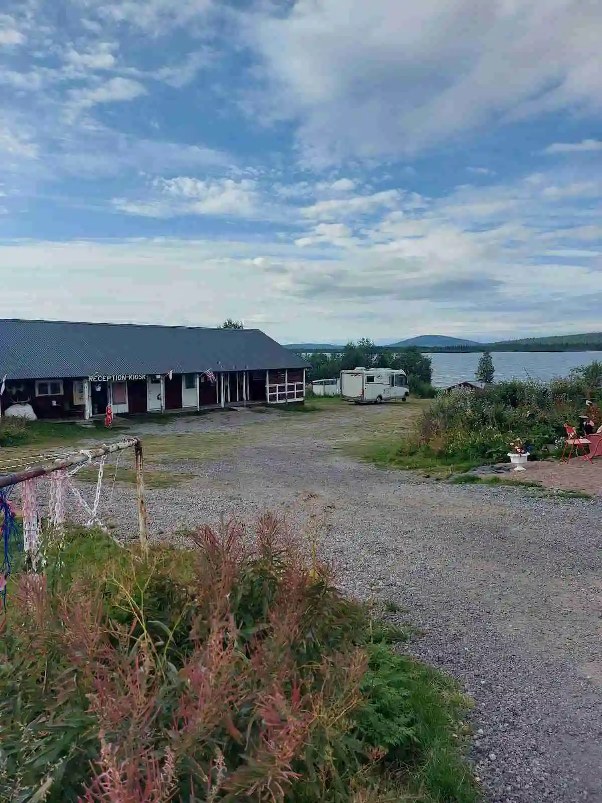 En klarblå himmel med några vita moln hänger ovanför Piilijärvi Camping. I förgrunden syns en gårdsplan där flera bilar, inklusive en lyxbil och en familjebil, står parkerade. Bakom bilarna finns en charmig stuga.