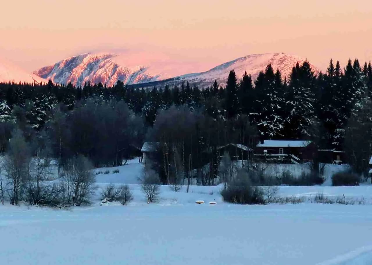 En vinterbild av Storsjö camping och ställplats med snötäckta berg och höjder i bakgrunden samt frostdraperade trädgrenar i förgrunden.
