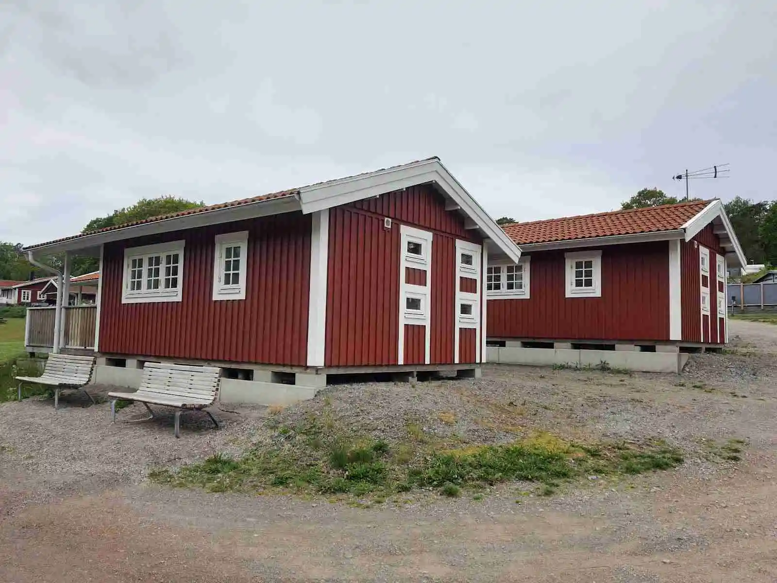 En trästuga med rött tak och vita knutar står på en grönskande campingplats. Stugan har en enkel dörr och är omgiven av ett gräsbevuxet område, vilket ger en lantlig och avkopplande atmosfär.