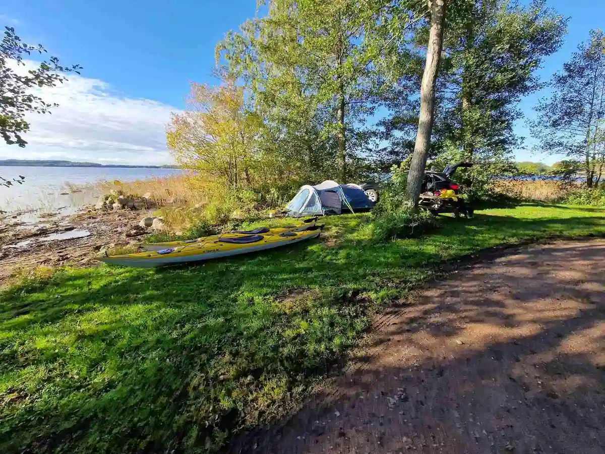 En campingplats vid kusten, med gröna växter i förgrunden och tält uppställda nära stranden. I bakgrunden syns en kajak och en båt vid vattnet.