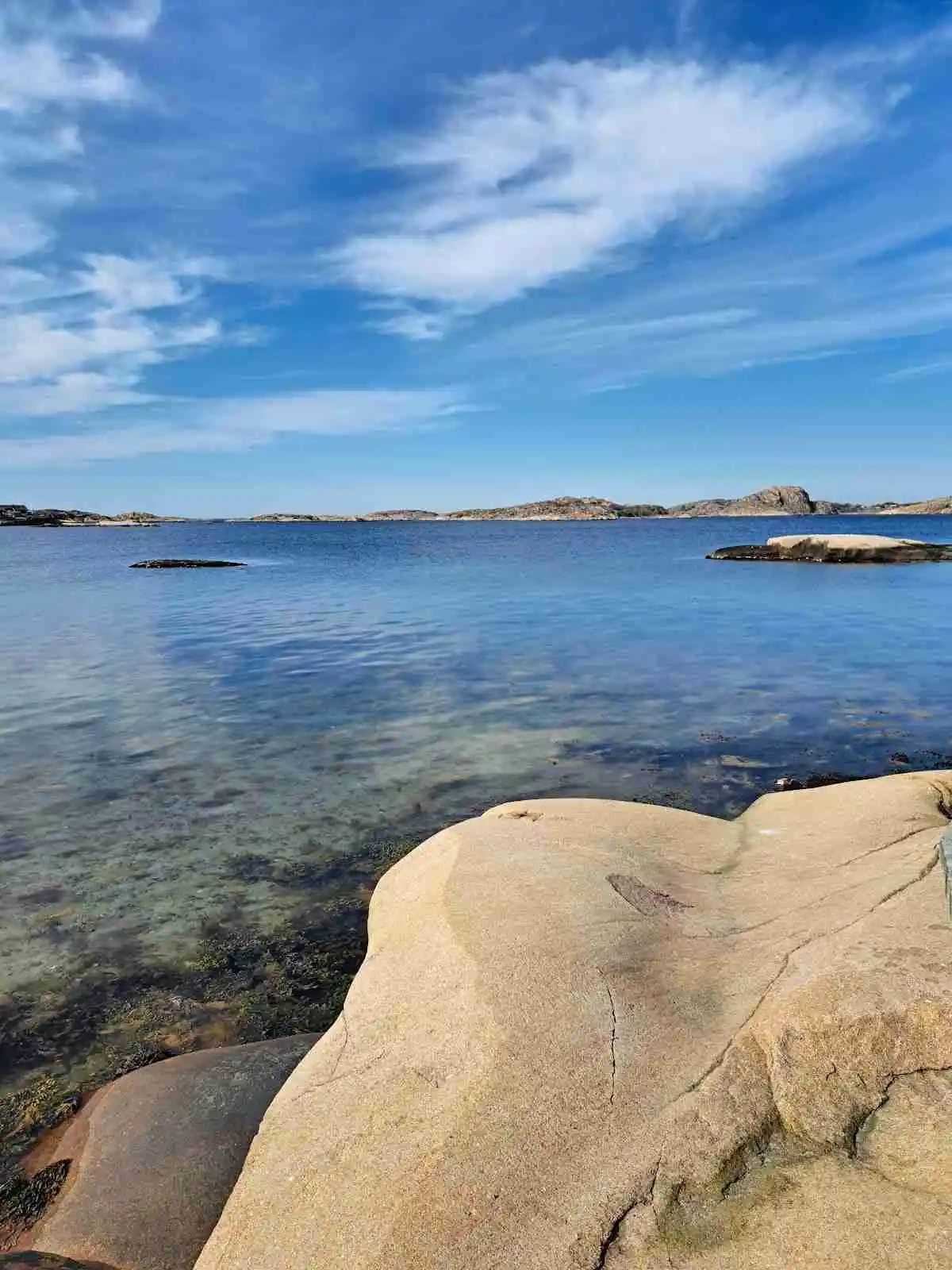 Utsikt över en solig kustlinje vid Hunnebostrands camping och stugor, med blått hav och klippor i förgrunden samt horisonten i bakgrunden.