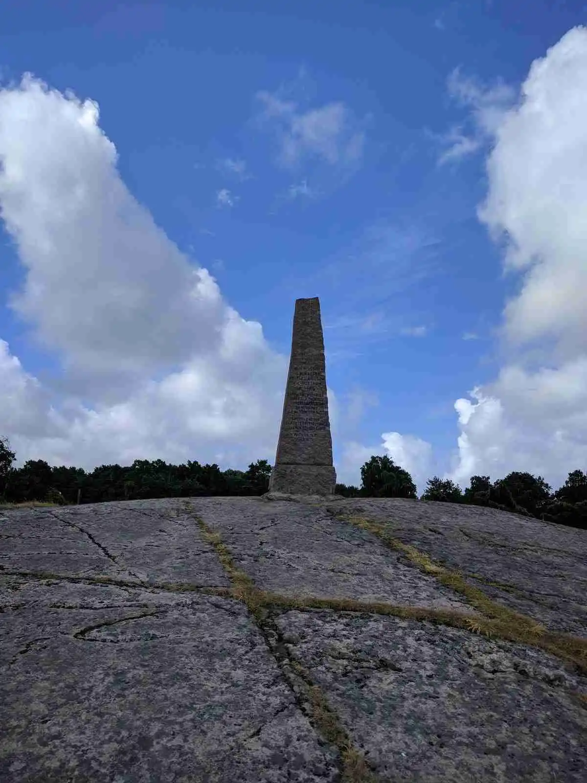 Alt-text: Ett monumentalt stenmonument reser sig i en naturskön miljö vid Vallersvik Camping and Hostel, omgivet av grönska och en klar himmel i bakgrunden.