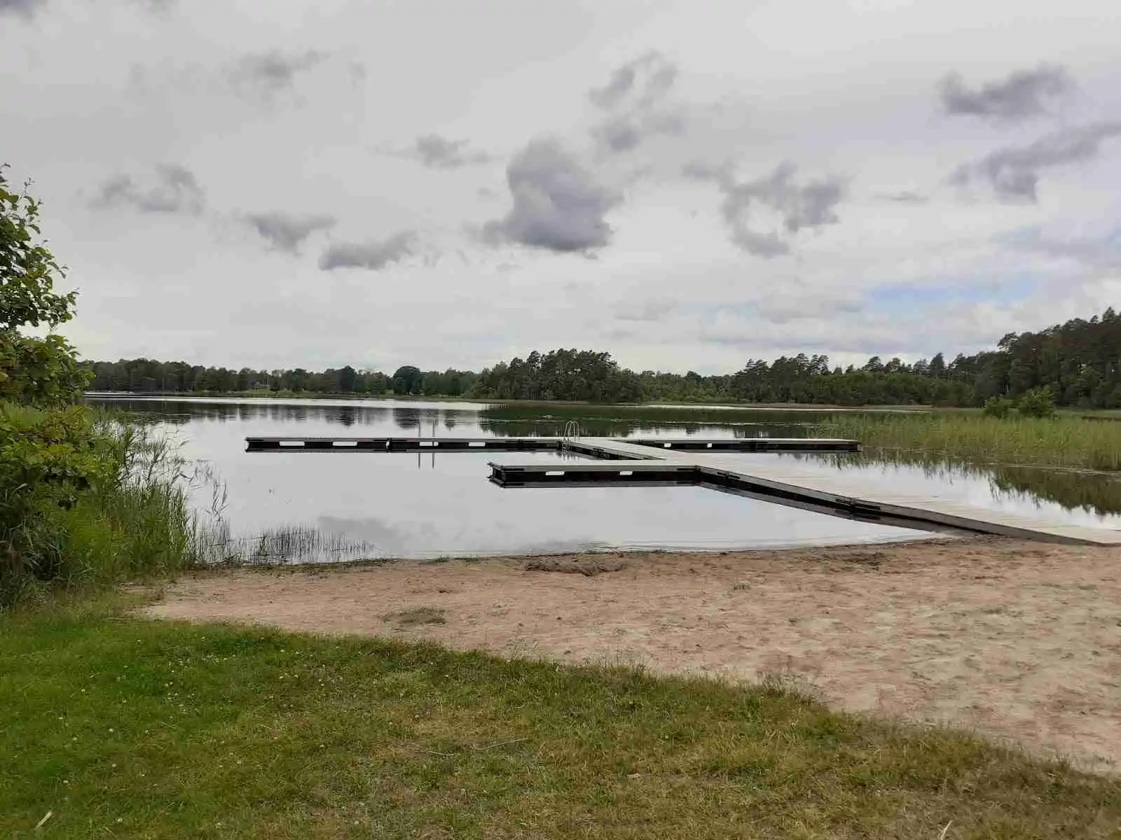 En naturskön vy över Linneryds camping med en sjö omgiven av grönskande strandbanker under en himmel med vita moln.