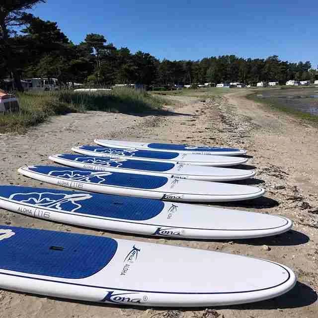 En bild av kyrkvikens camping ab med fokus på vattensporter. En surfbräda och annan utrustning ligger vid vattnet, redo för aktiviteter som surfing och båtliv. Solen skiner och skapar en atmosfär av avkoppling och nöje vid sjön.