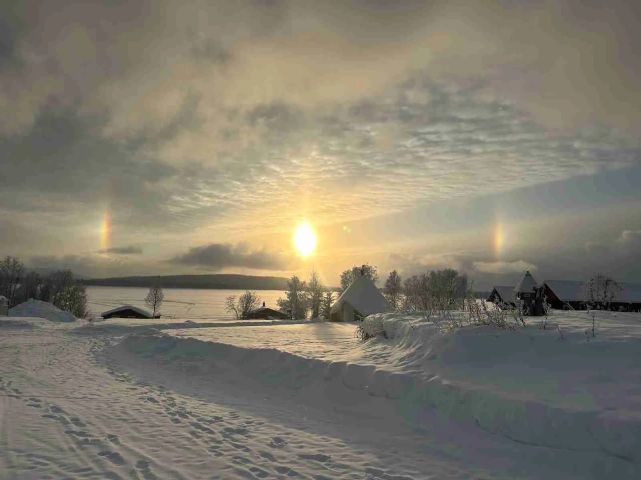 En snötäckt vinterlandskap med en uppgående sol som färgar himlen i varma nyanser. Fluffiga moln och en klar himmel skapar en fridfull och stillsam atmosfär över Piilijärvi camping.