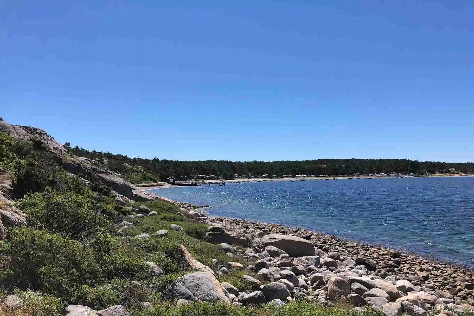 Vy över Kyrkvikens Camping AB med blått vatten och kustlinje. Stenar syns längs strandkanten under en klar sommarhimmel.