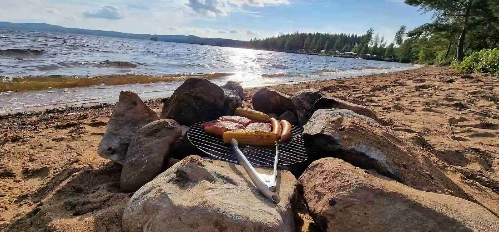 En grill står ute på en naturcamping med grillgaller över glödande kol. Omkring finns utomhusmöbler som bidrar till campingstämningen.
