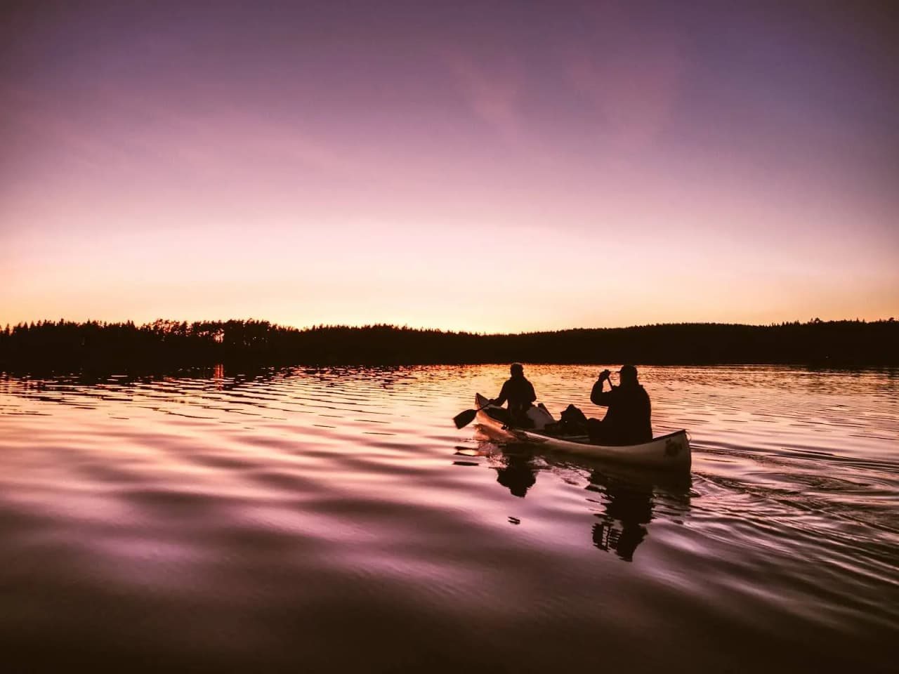 En båt ligger förtöjd vid sjön i skymningen, omgiven av stilla vatten och reflekterande solnedgångsljus på Viking Republic Nature Camp.