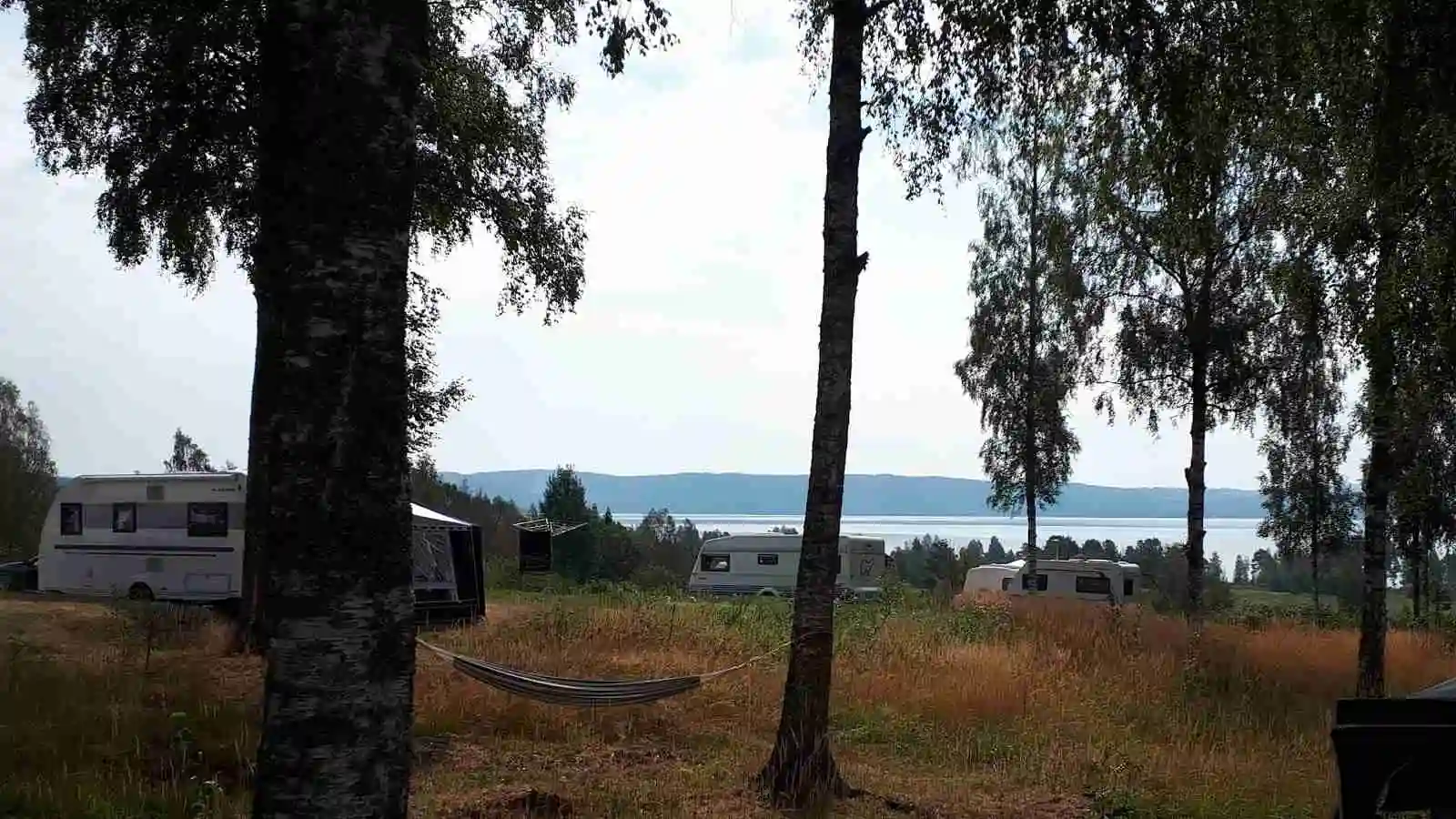 En husvagn parkerad på en grusplan vid Vikarbygården, omgivet av grönska och en klar himmel.