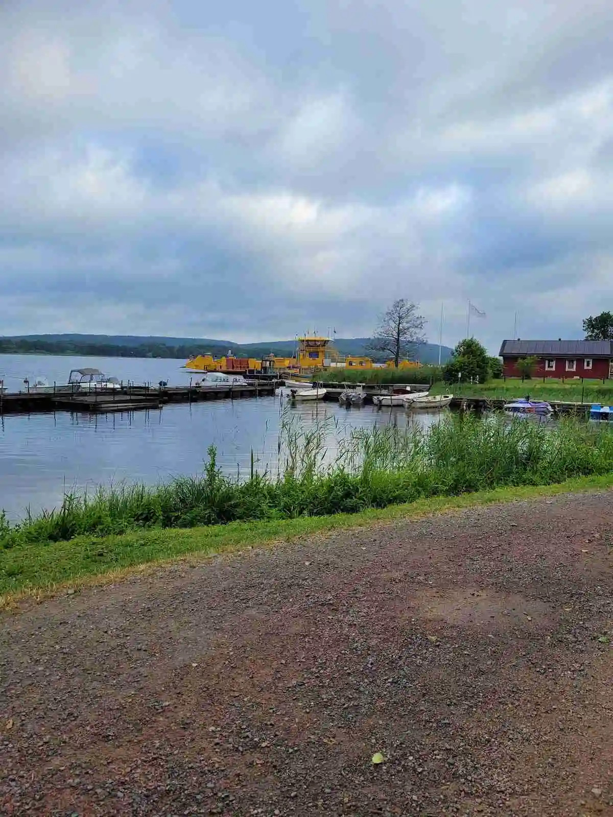 En solig dag vid Ivö Camping med moln på himlen och utsikt över en sjö. Flera båtar ligger förtöjda vid en brygga längs kusten.