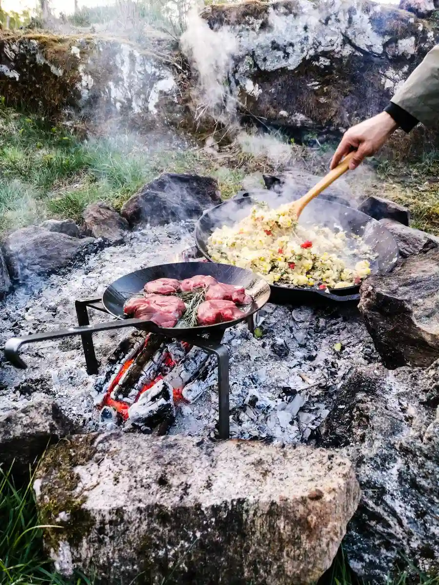 Ett utomhuskök på Viking Republic Nature Camp där kött grillas över öppen eld. Eldslågor och rök syns medan maten tillagas på en robust grillplats, omgiven av naturen.