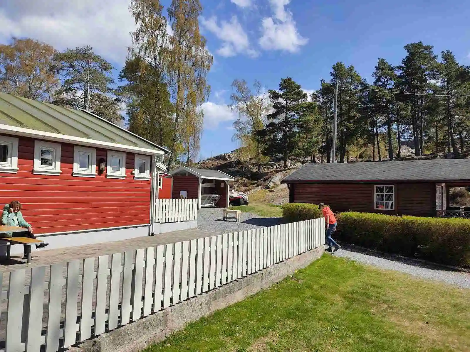En lantlig miljö med ett charmigt rött hus med vita knutar, omgiven av grönska och buskar, typiskt för en svensk landsbygdsboende med en trivsam och hemtrevlig atmosfär.